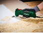 Green multi-tool with a gloved hand scraping adhesive from a wooden surface.