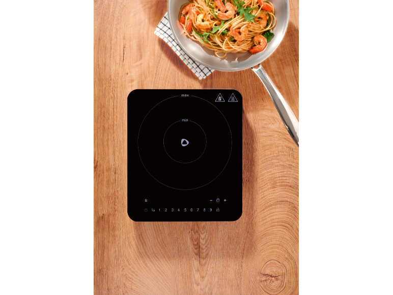 Induction hob with pasta with shrimp and arugula in a pan on a wooden table.