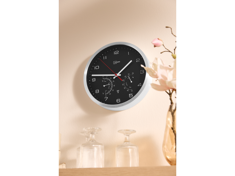 Wall clock with thermometer and hygrometer on a light beige wall, with a vase of flowers.