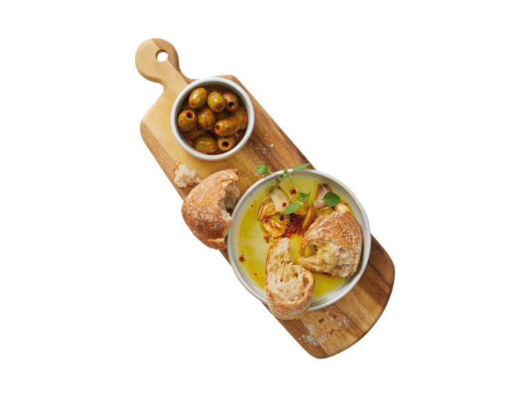 Wooden board with green olives, bread, and a bowl of olive oil with garlic and herbs.