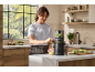 Smiling woman using a black juicer to make green fruit juice in a modern kitchen.