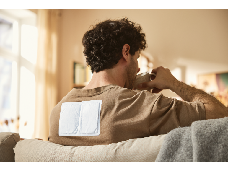 Man with warming patches on his back drinks coffee on the couch.