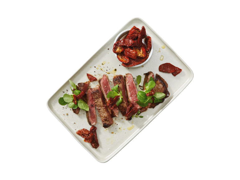 Sliced steak with sun-dried tomatoes and green leaves on a rectangular platter.