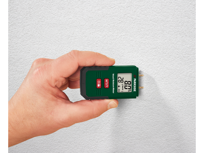 Hand holding a wood moisture meter against a white textured wall, displaying 28°C and 8.0%.