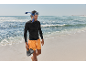 Man with a snorkel mask, black long-sleeve shirt, and orange shorts walks on the beach.