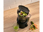 Black juicer with chopped vegetables and a glass of green juice on a wooden countertop.