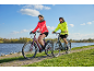 Two people in brightly colored cycling jackets ride bicycles on a path by a river.