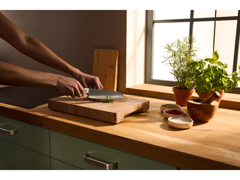 Hands cutting herbs on a wooden chopping board, next to spices and potted herbs.