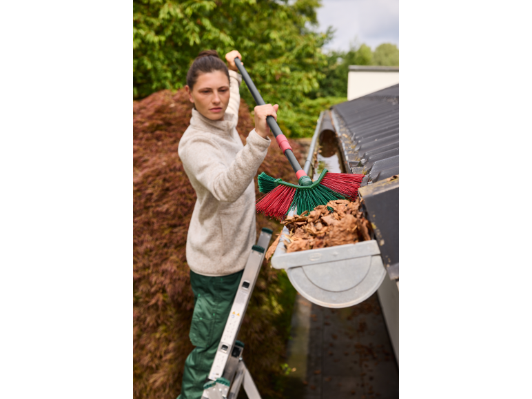 Woman cleaning a gutter with a telescopic brush, standing on a ladder.