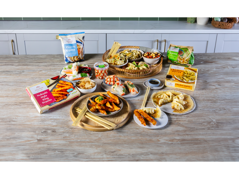 A wooden table with a variety of Japanese-style products, including prawns, gyoza, and spring rolls.