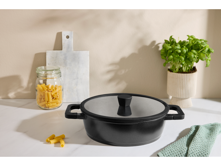 Black pot with lid, jar of pasta, cutting board, and basil plant on a countertop.