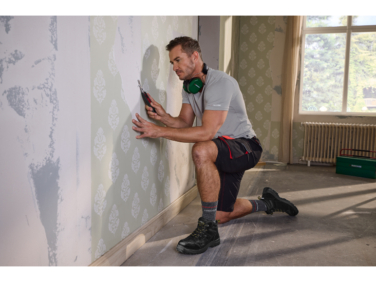 Man in shorts and grey t-shirt, kneeling, removing wallpaper with a scraper.