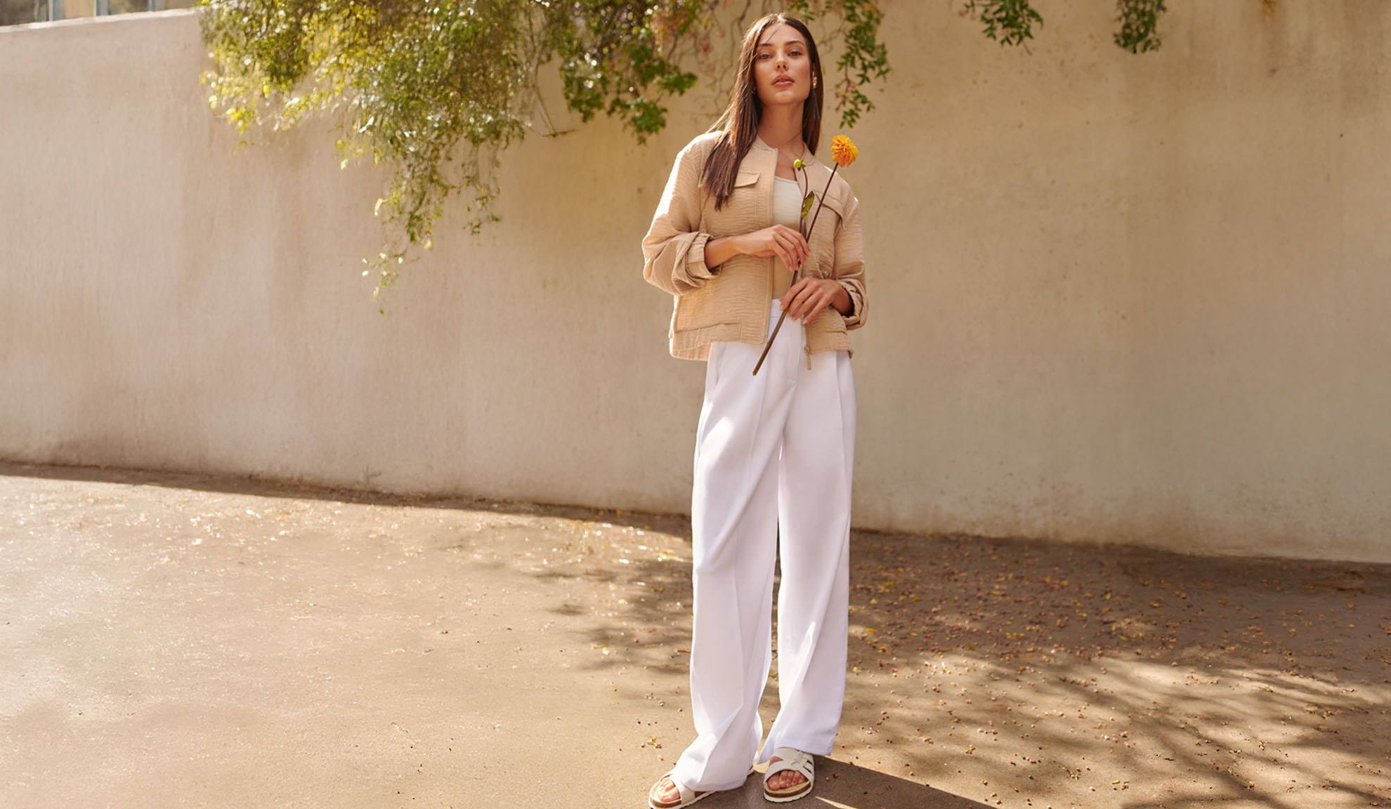 Woman in beige jacket, white trousers, and sandals, holding a yellow flower.