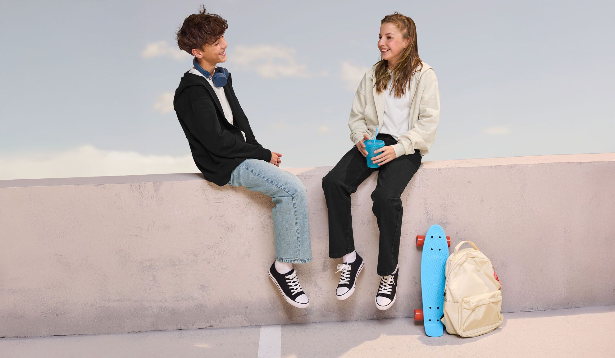 Two teenagers in casual wear, sitting on a wall, with a skateboard and backpack nearby.