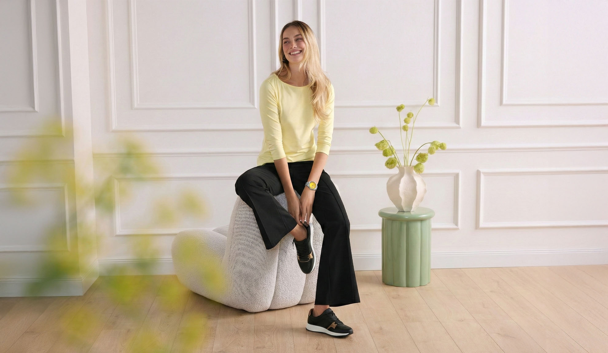 Woman in yellow long-sleeve top and black trousers, sitting on a pouf, wearing sneakers.