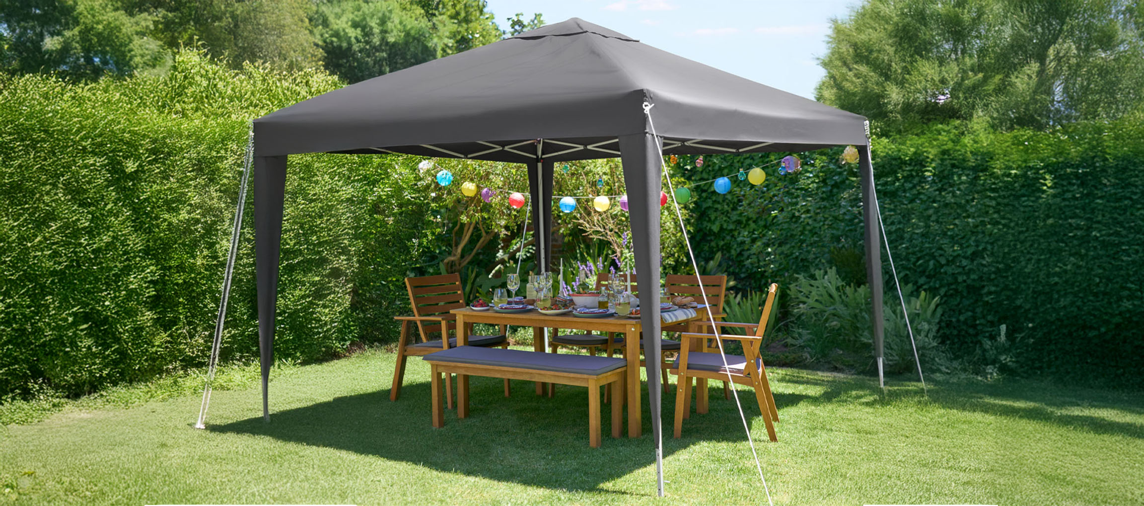 A dark gray pop-up gazebo shelters a wooden outdoor dining set in a lush green garden.