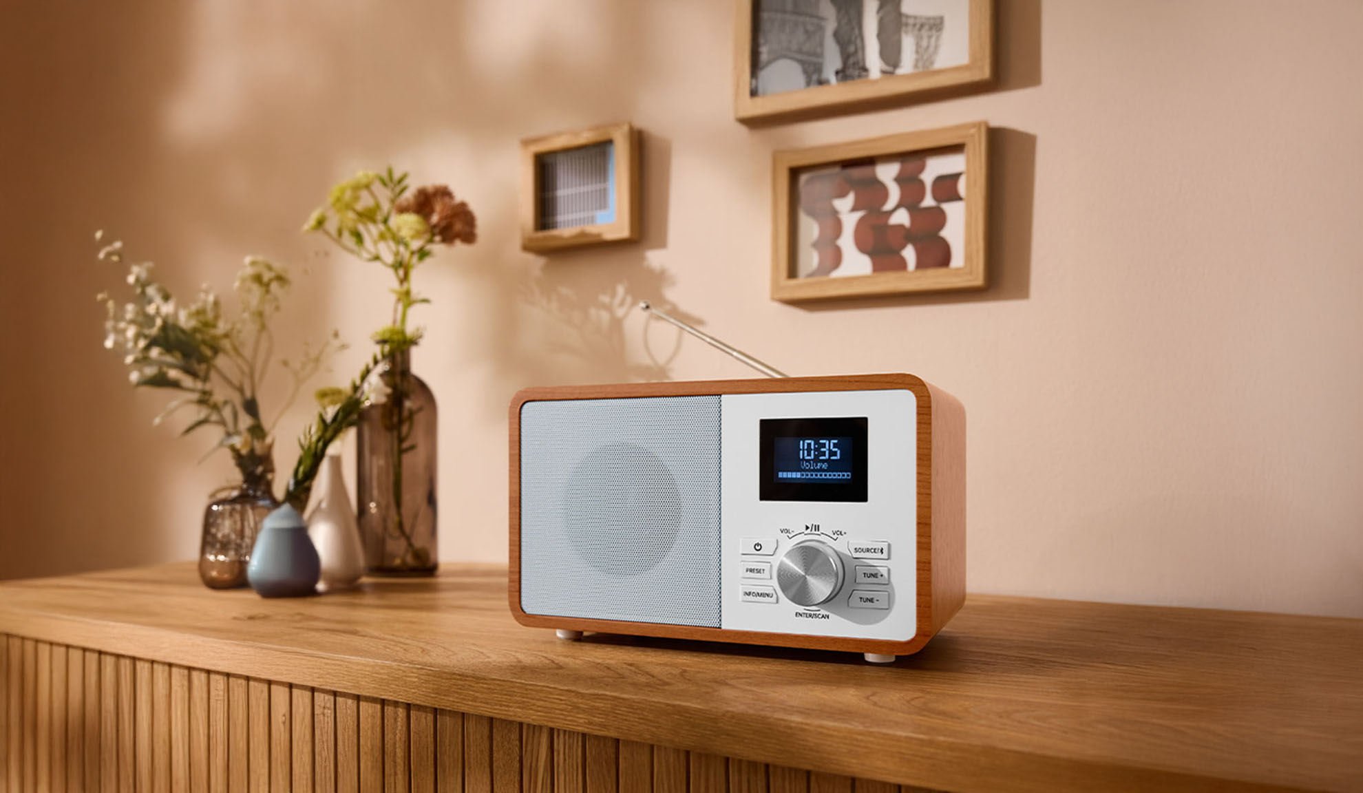 A wooden digital radio on a dresser, with vases and picture frames in the background.