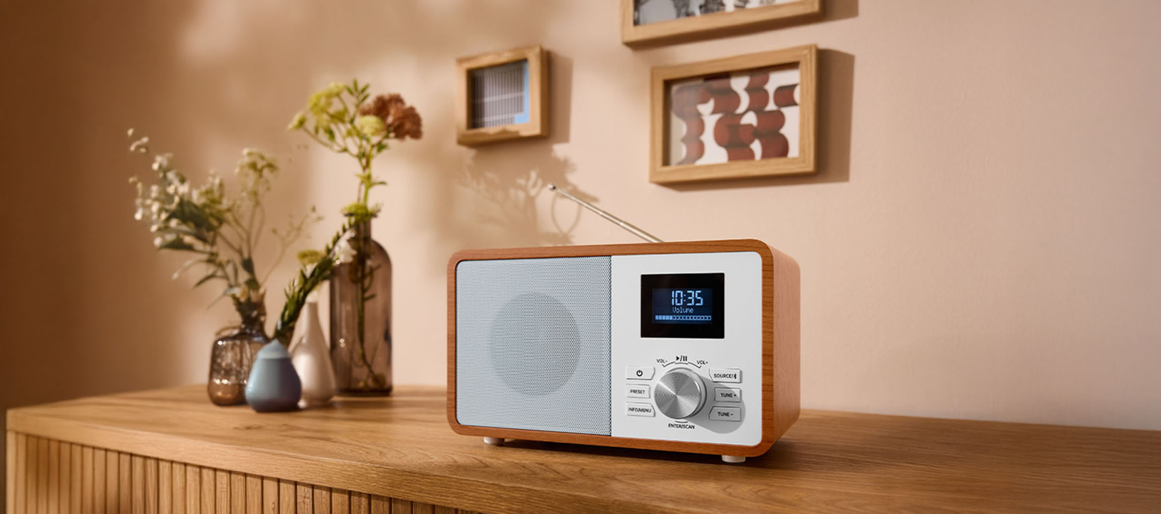 A wooden digital radio on a dresser, with vases and pictures on the wall.