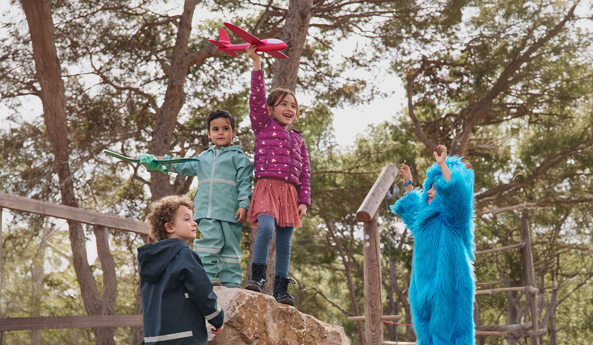 Four children playing outdoors, two with toy airplanes and one in a blue monster costume.