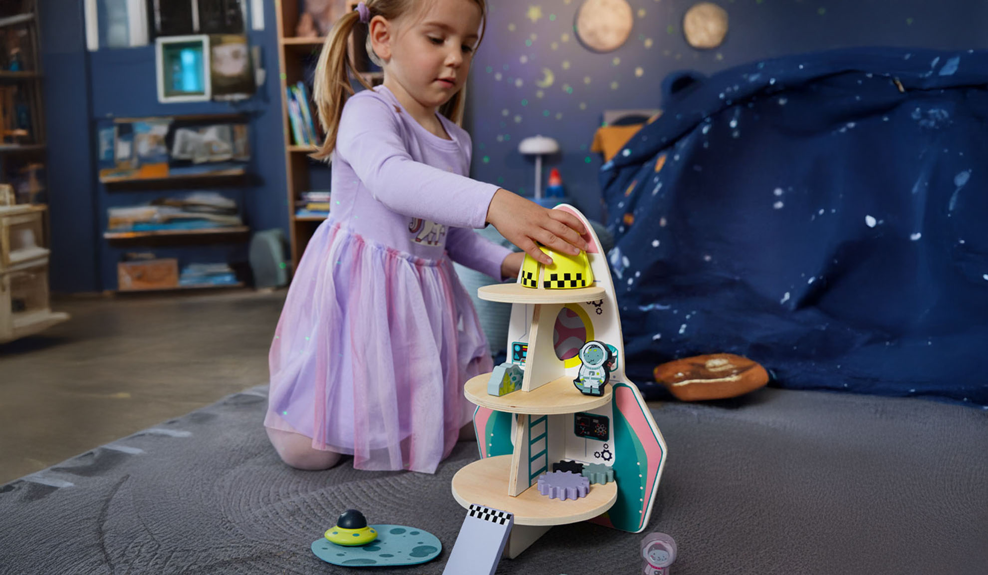 A girl plays with a toy rocket and space accessories in a themed room.
