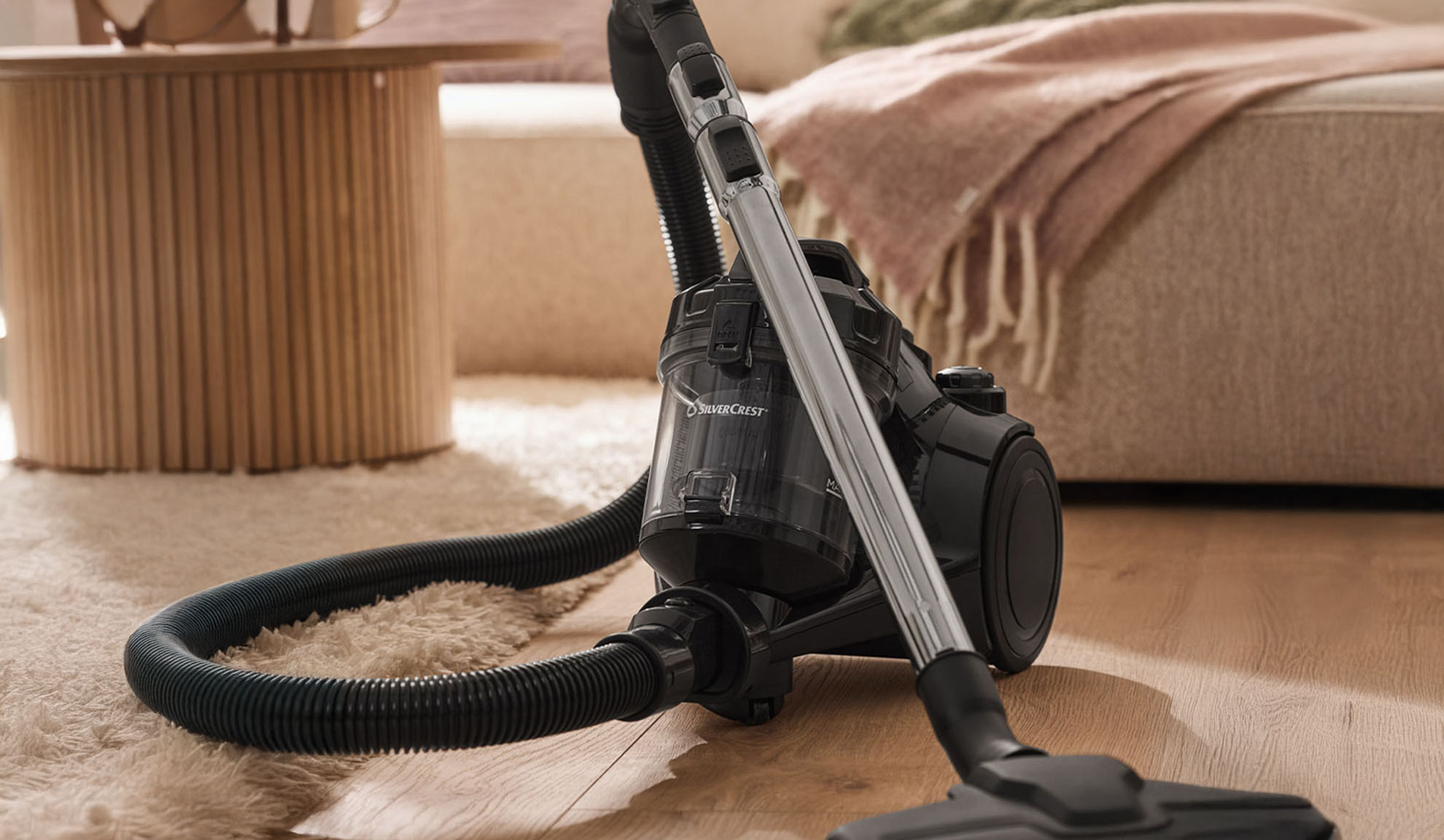 Black vacuum cleaner on a wooden floor, with a fluffy rug and furniture in the background.