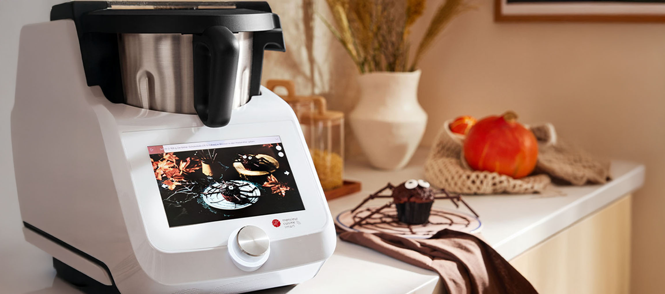 Kitchen robot with a touchscreen displaying a Halloween recipe, next to a spider cupcake.