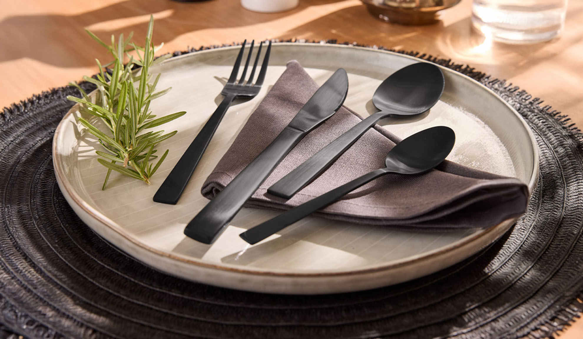 Black cutlery set with a plate, napkin, and rosemary sprig on a placemat.