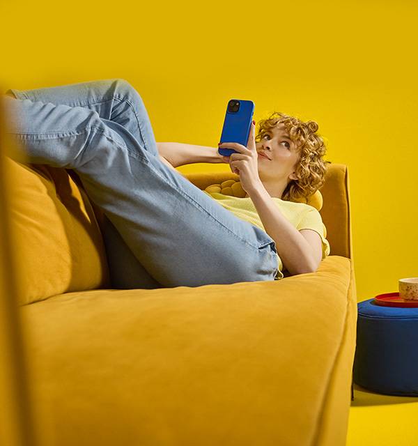 Woman on a yellow sofa using a blue smartphone, with a blue stool nearby.