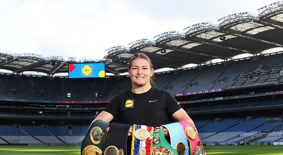 Female boxer holding championship belts in a stadium, with a large screen in the background.