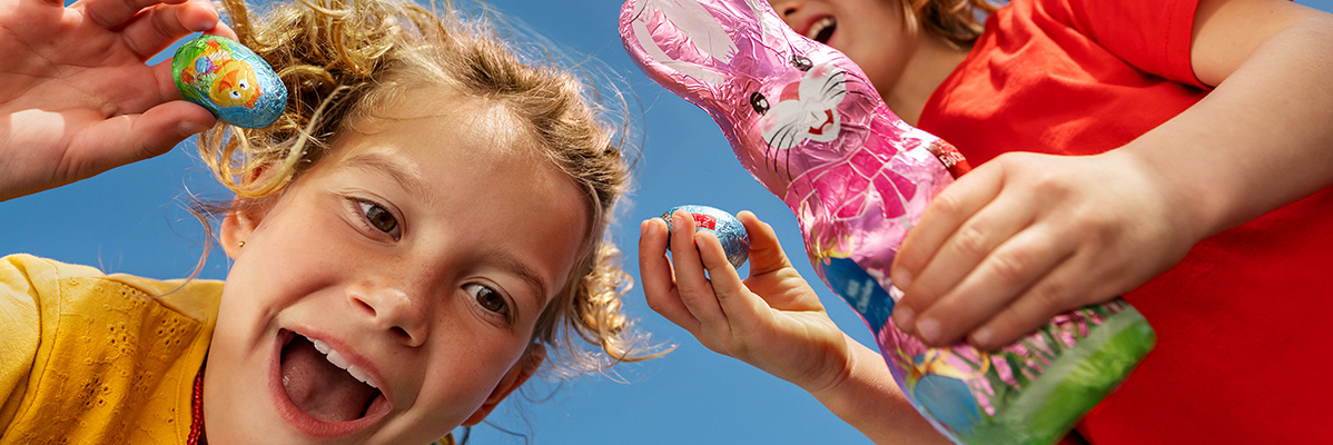 Two happy children holding chocolate Easter eggs and a chocolate bunny against a blue sky.
