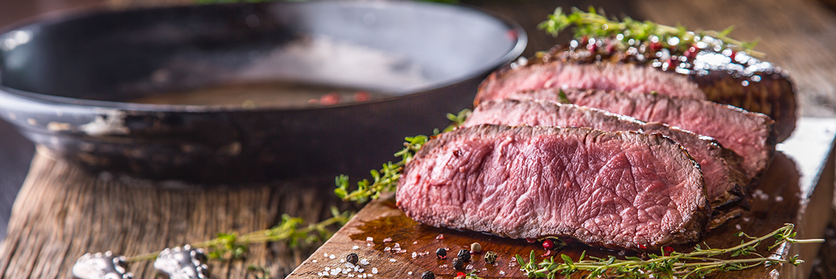 Sliced medium-rare steak with herbs and spices on a wooden board, with a pan in the background.