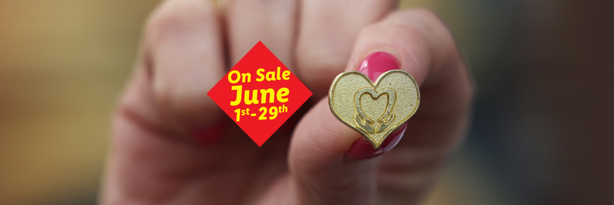 A hand holding a golden heart-shaped pin, with a red sale tag for June 1st-29th.