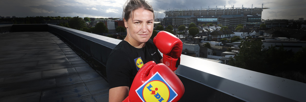 Female boxer in black t-shirt and red boxing gloves with a stadium in the background.