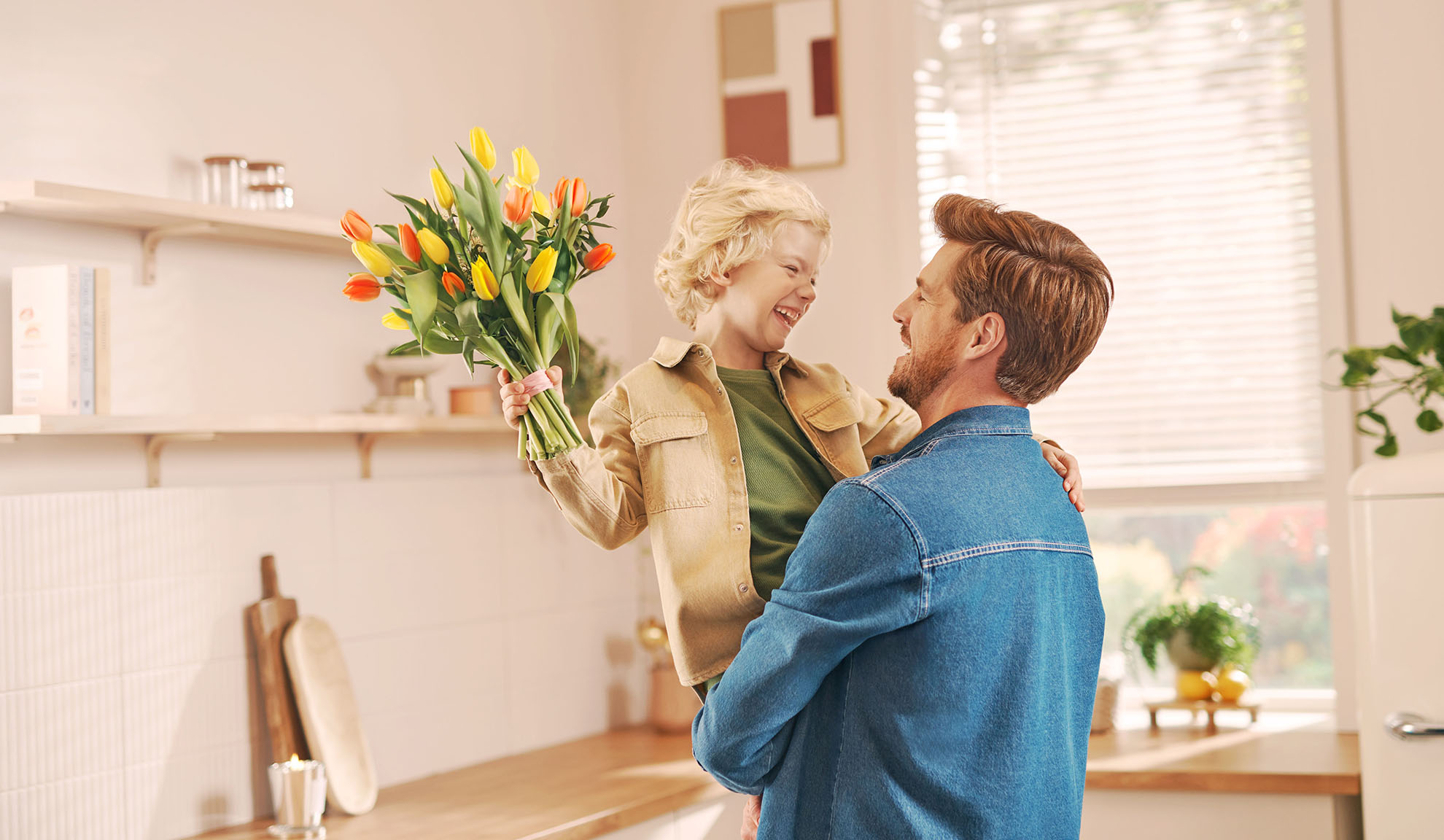 Father holding son who is presenting a bouquet of tulips in a bright kitchen.