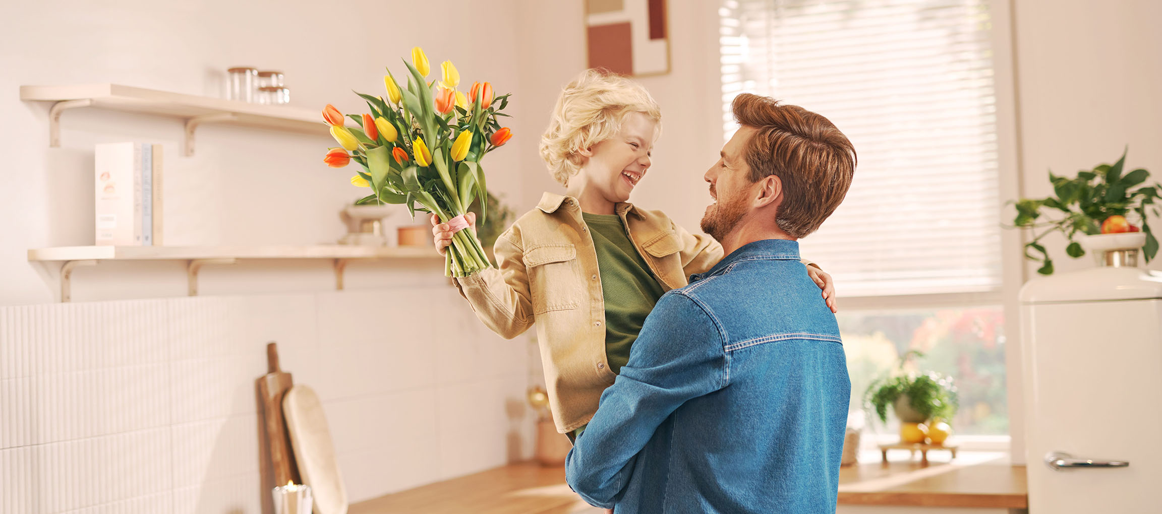 A boy holding a bouquet of tulips, laughing with a man in a kitchen.