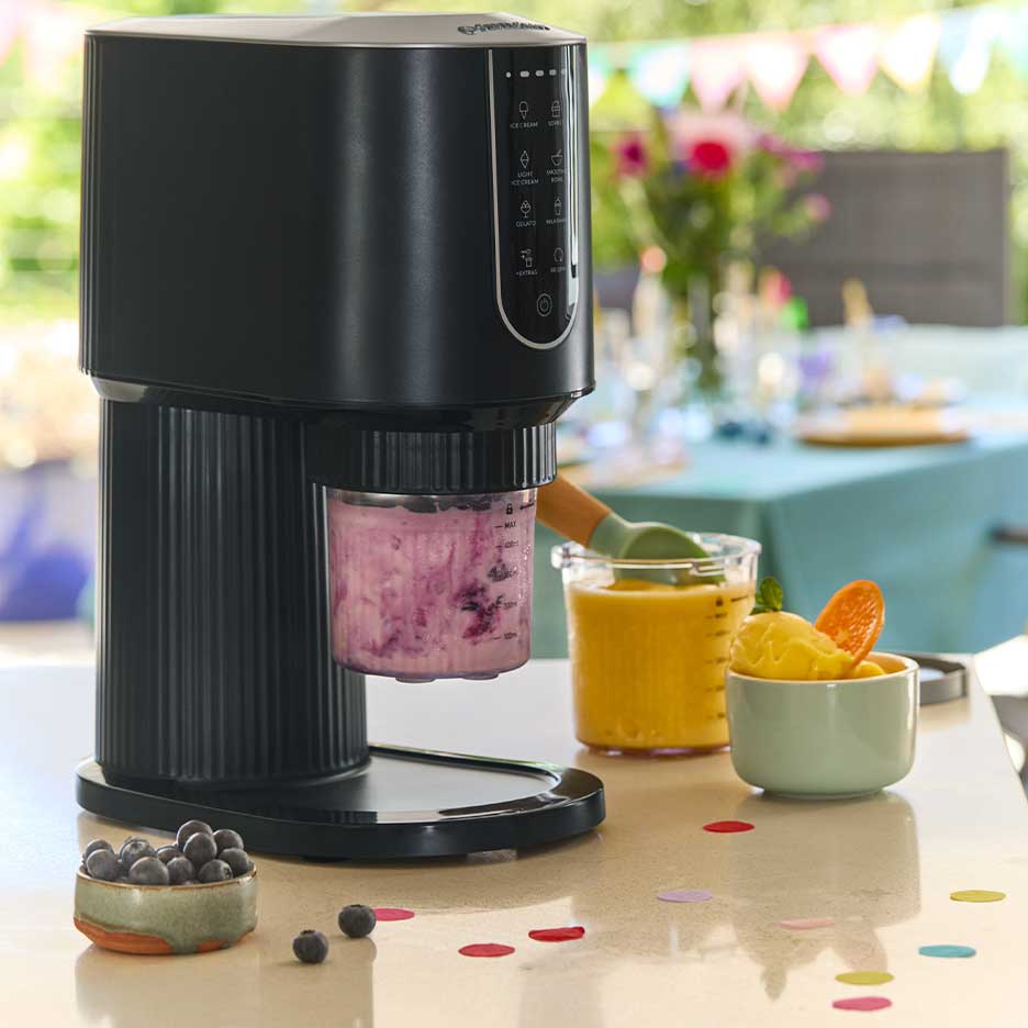 Black ice cream maker with purple ice cream, surrounded by mango ice cream and blueberries.