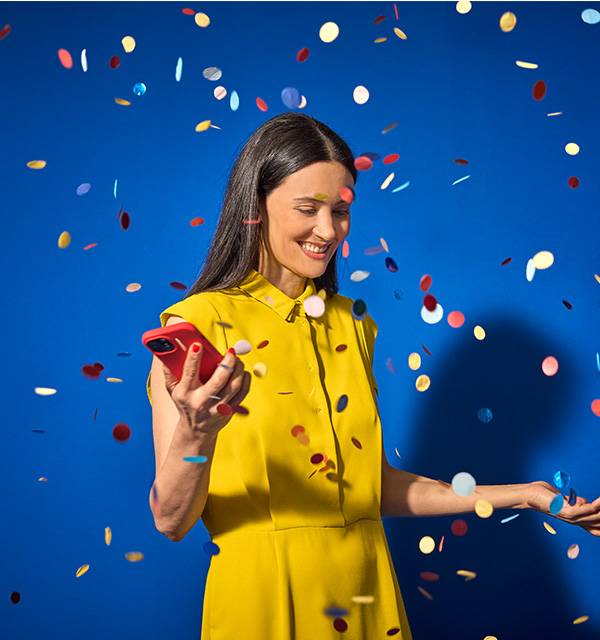 Woman in yellow dress holding a red smartphone, smiling amidst falling confetti.