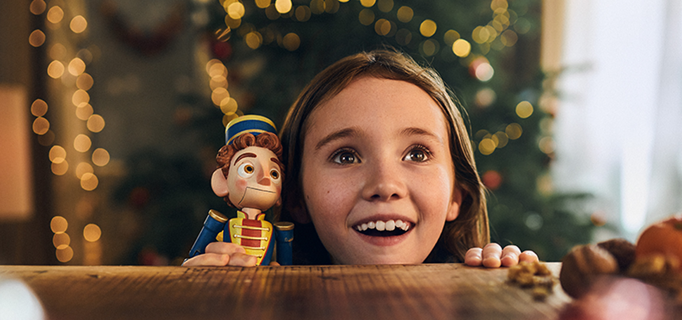 A smiling young girl peeking over a table next to a toy nutcracker with Christmas lights behind.