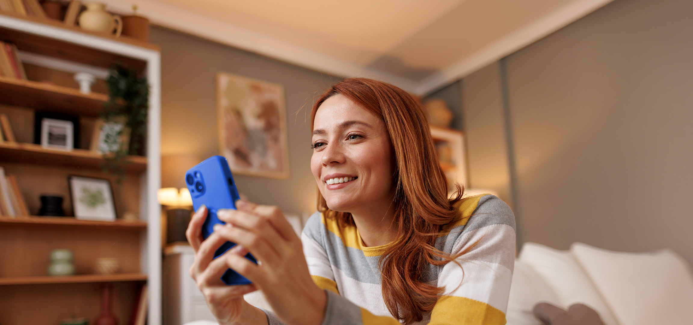 Smiling woman with red hair holding a blue smartphone in a cozy living room.