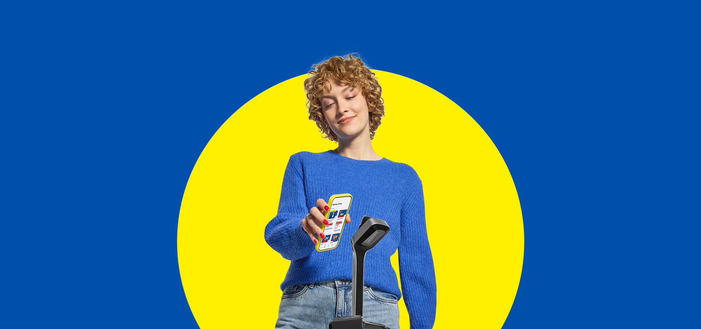 A young woman in a blue sweater scanning a phone with a mobile app at a self-checkout.