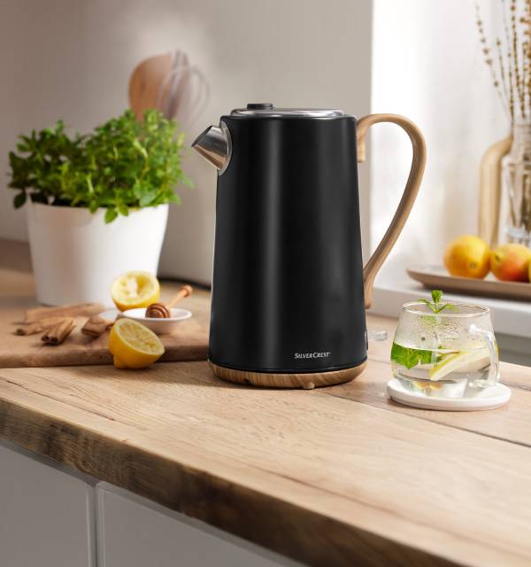 Black kettle with wooden handle on a kitchen counter with lemons and herbs.