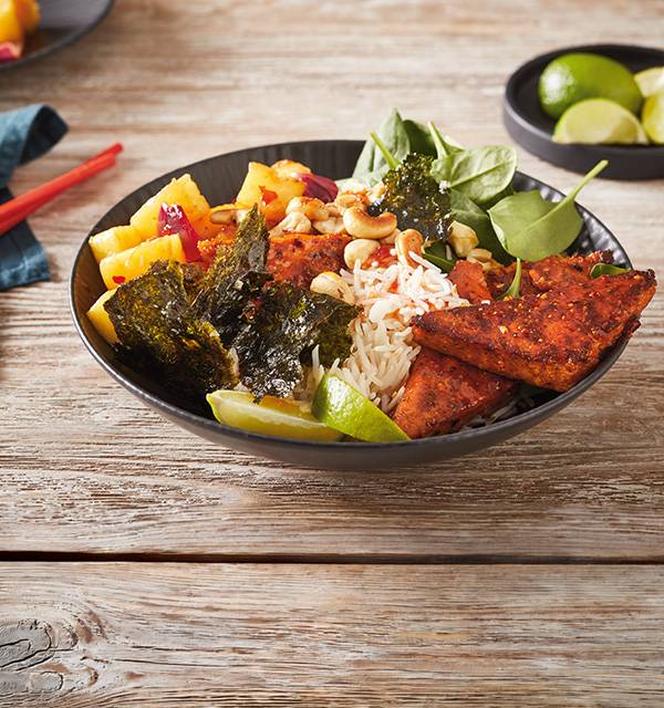 A colorful poke bowl with rice, tofu, pineapple, spinach, seaweed, cashews, and lime wedges.