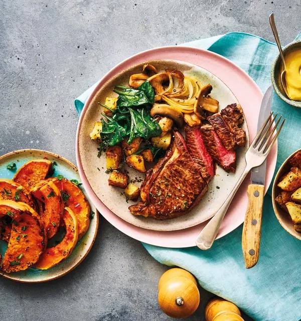 Steak with roasted potatoes, spinach, mushrooms, and butternut squash on a grey background.