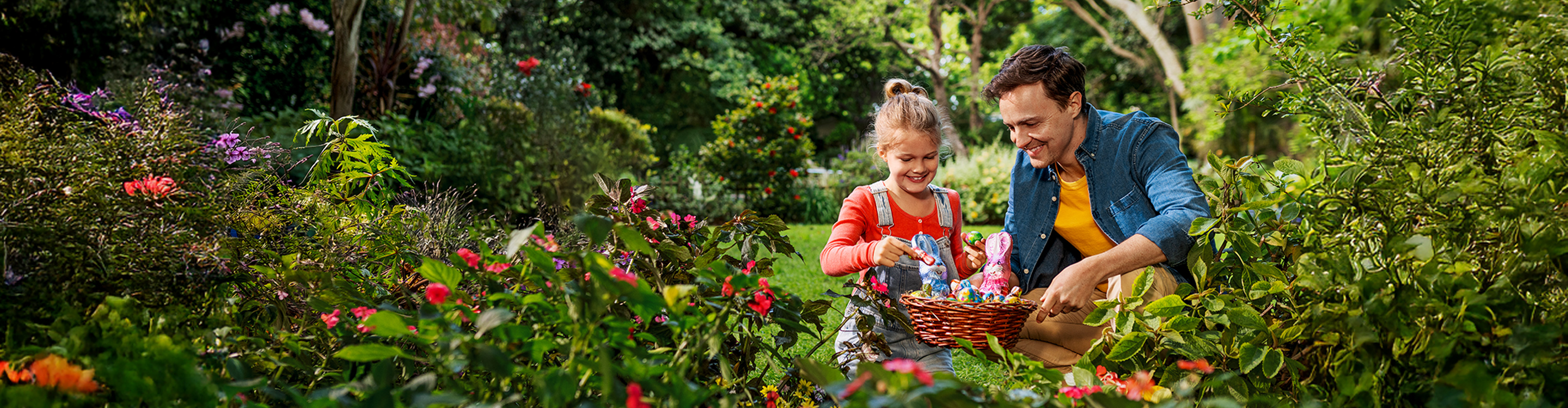 Father and daughter with Easter basket filled with chocolate bunnies and eggs in a garden.