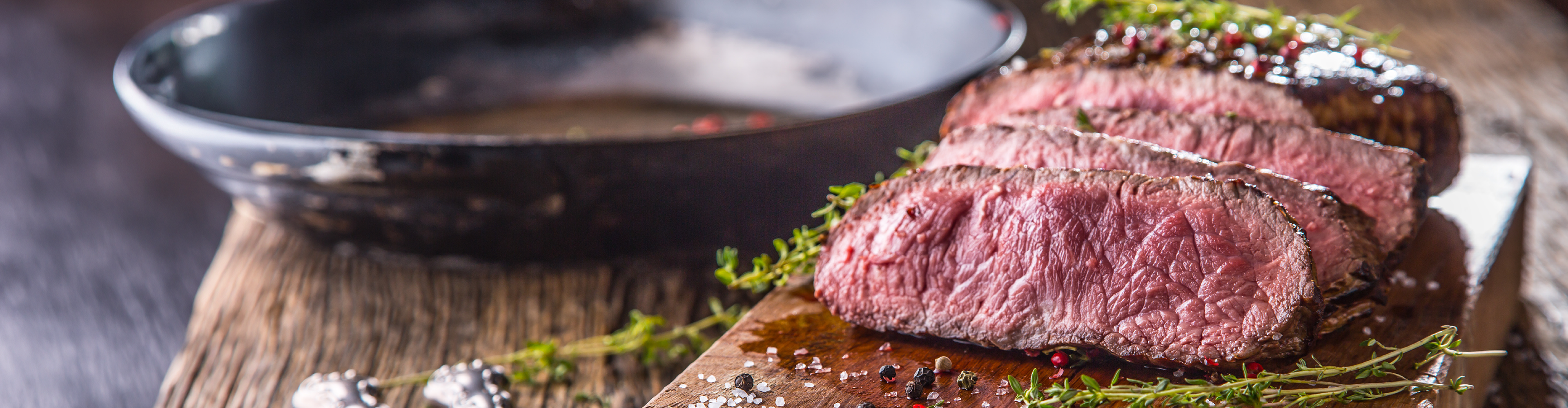 Sliced medium-rare steak with herbs and spices on a wooden board, with a pan in the background.