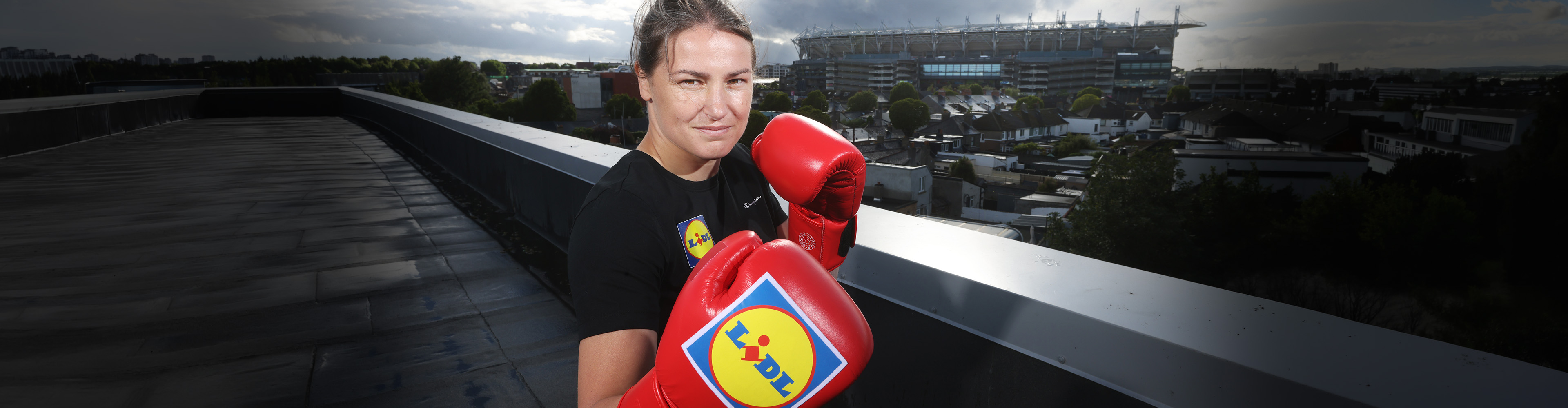 Female boxer in black t-shirt and red boxing gloves with a stadium in the background.