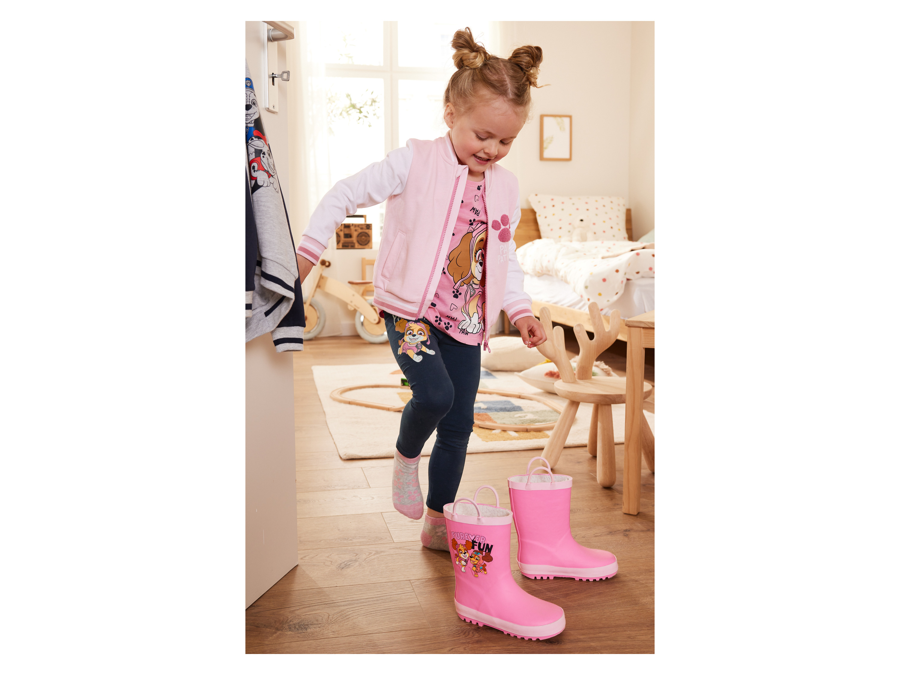 A toddler girl tries on pink Paw Patrol rain boots in a bedroom.
