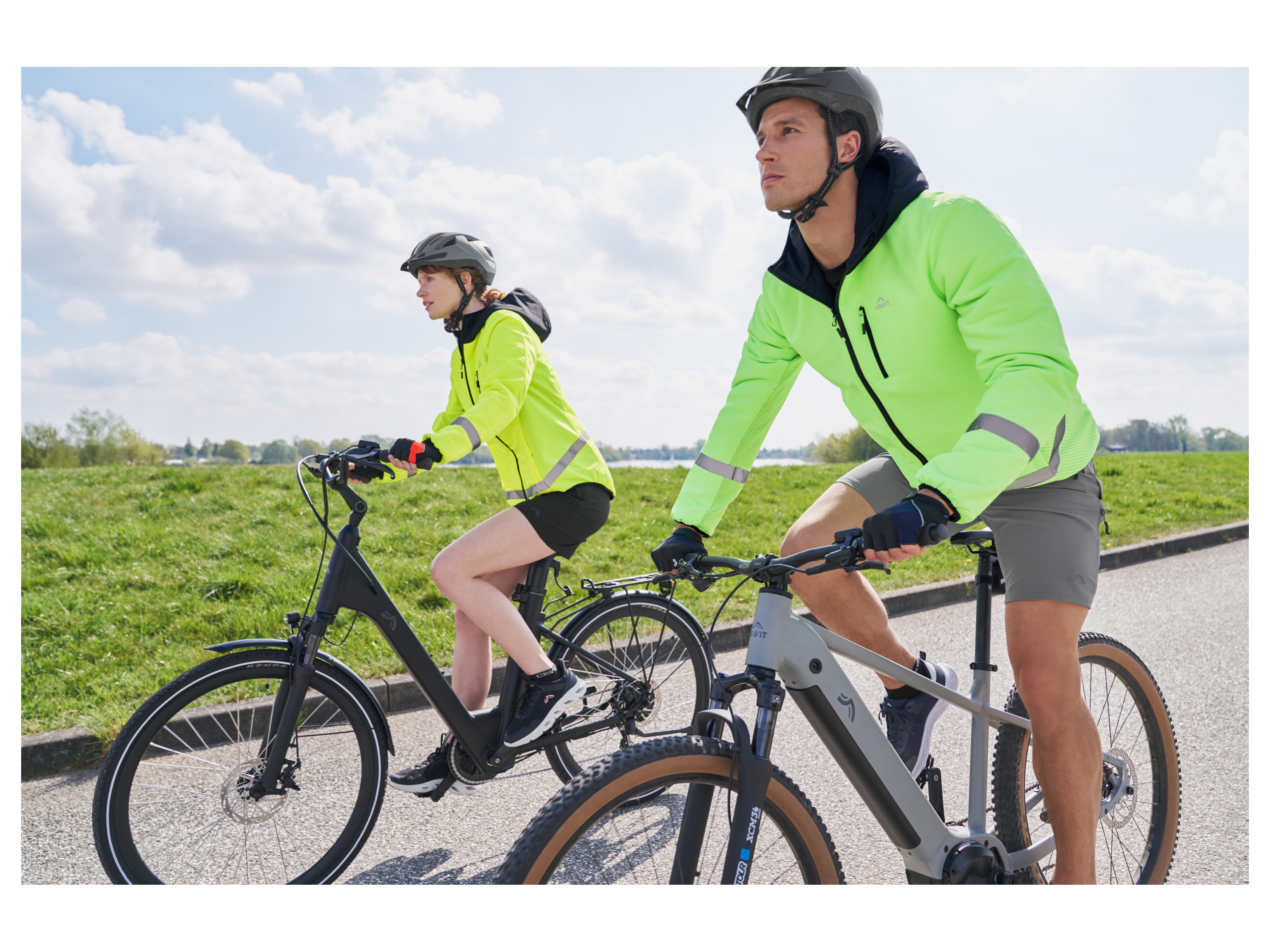 Two people ride electric bikes wearing bright cycling jackets and helmets on a sunny day.