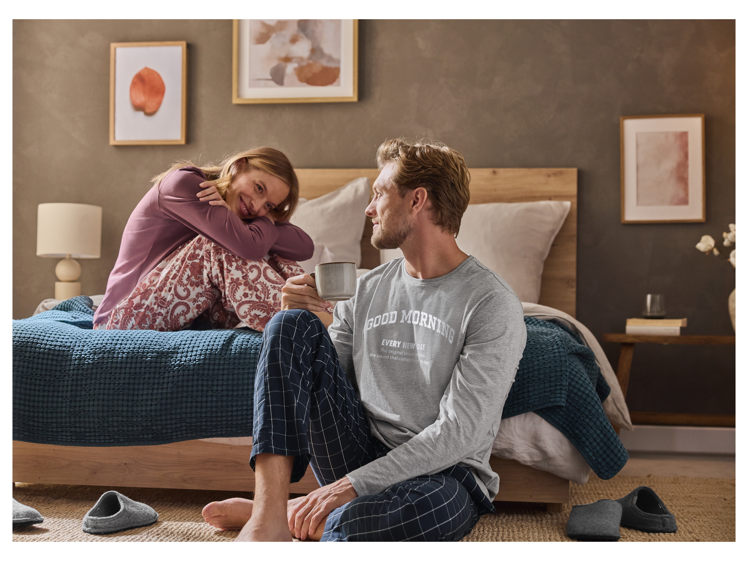 A couple in pajamas, the man holding a mug, sitting on the floor and bed in a bedroom.
