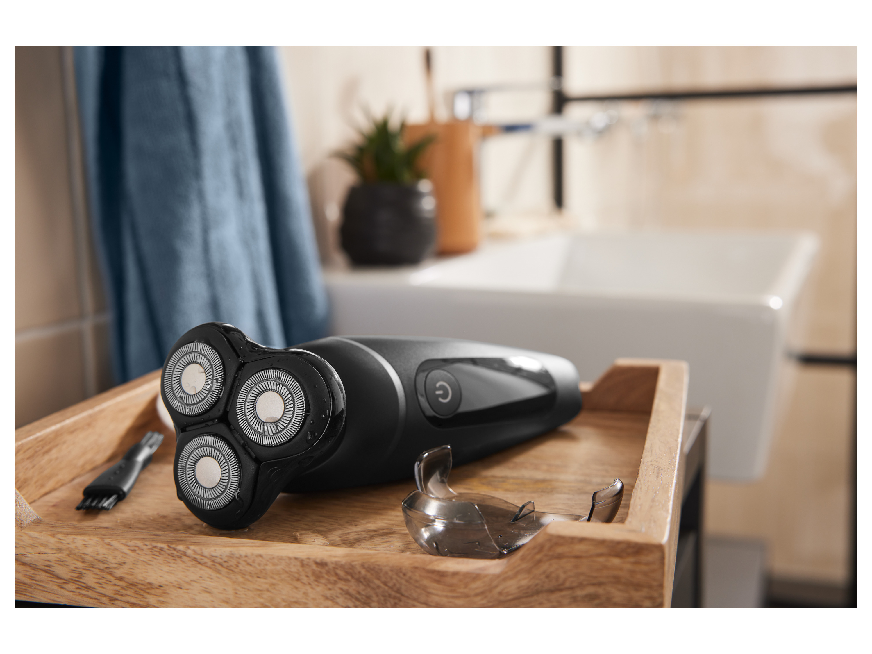 A black electric razor and its protective cap rest on a wooden tray in a bathroom setting.